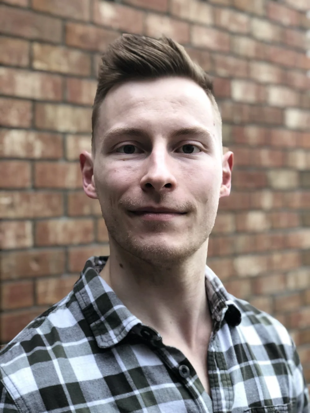 A young man with short brown hair, wearing a black and white checkered shirt, standing outdoors in front of a red brick wall.