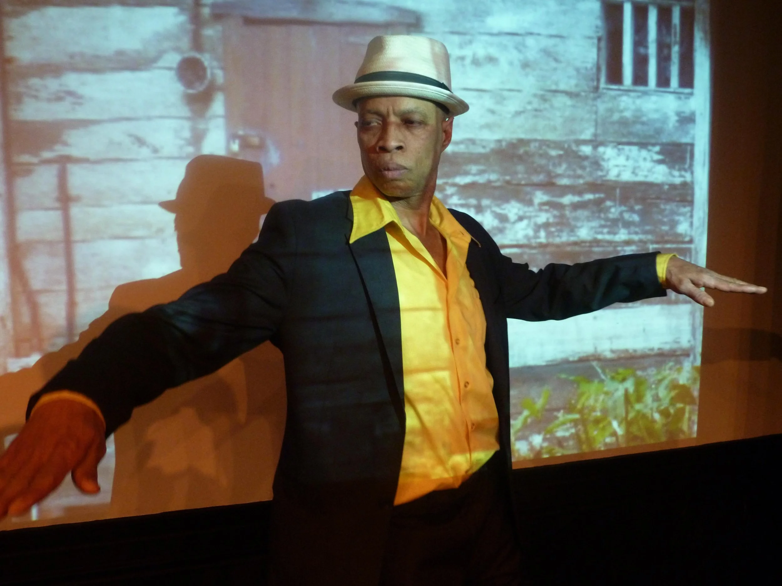 A man in a yellow shirt and black blazer wearing a white fedora hat stands with arms extended in front of a projected image of a rustic wooden house exterior.