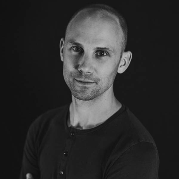 Black and white portrait of a man with short hair and a slight smile, wearing a buttoned shirt, against a dark background.