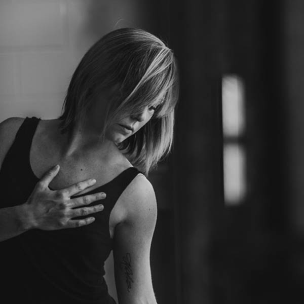A woman with shoulder-length hair touching her chest with her right hand, looking down thoughtfully.