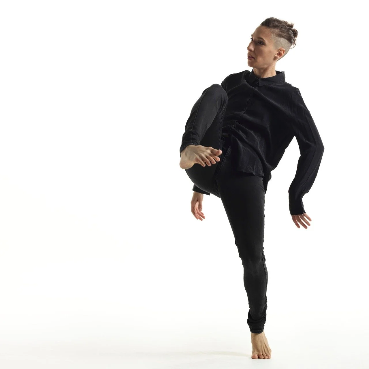 A woman with a mohawk hairstyle dressed in black, wearing a black shirt and pants, standing on one leg with the other leg raised and bent at the knee, balancing on her toes in a martial arts pose.