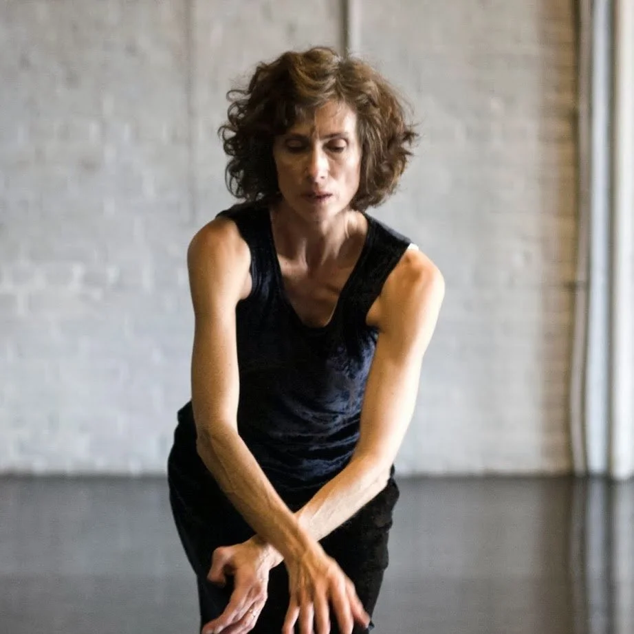 A woman with curly brown hair wearing a black sleeveless top and black pants standing in a dance studio with a gray brick wall background.
