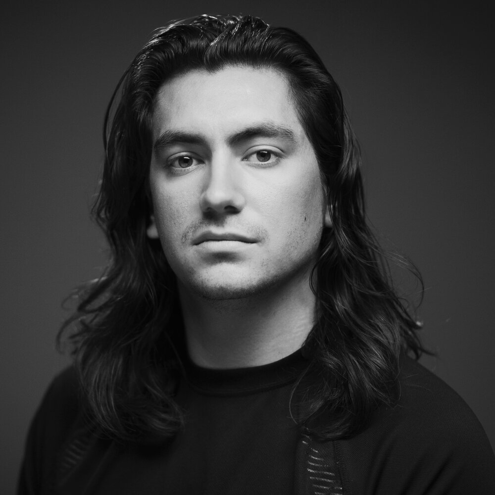 Black and white portrait of a young man with long wavy hair, wearing a dark shirt, looking directly at the camera with a neutral expression.