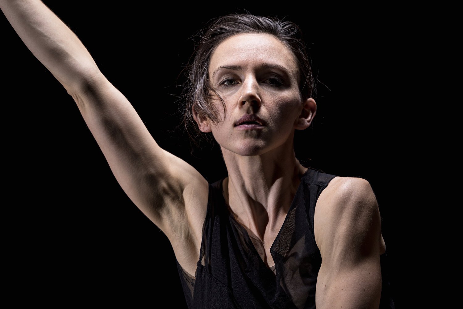 A woman with short dark hair, wearing a black sleeveless top, raising her right arm with a serious expression against a black background.