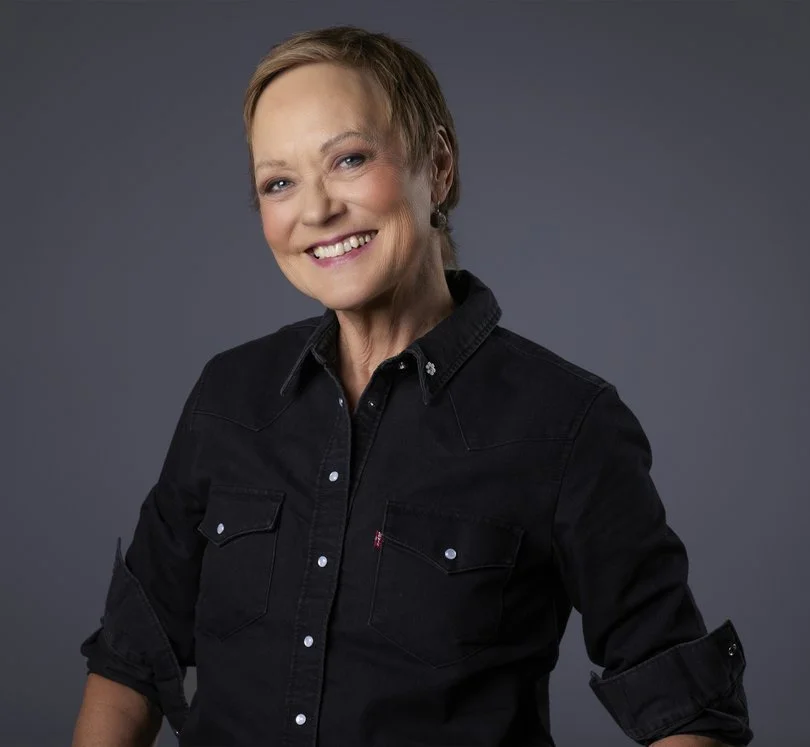 A smiling woman with short hair wearing a black denim shirt against a gray background.