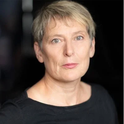 A woman with short blonde hair and blue eyes, wearing a black top, looking directly at the camera with a neutral expression.
