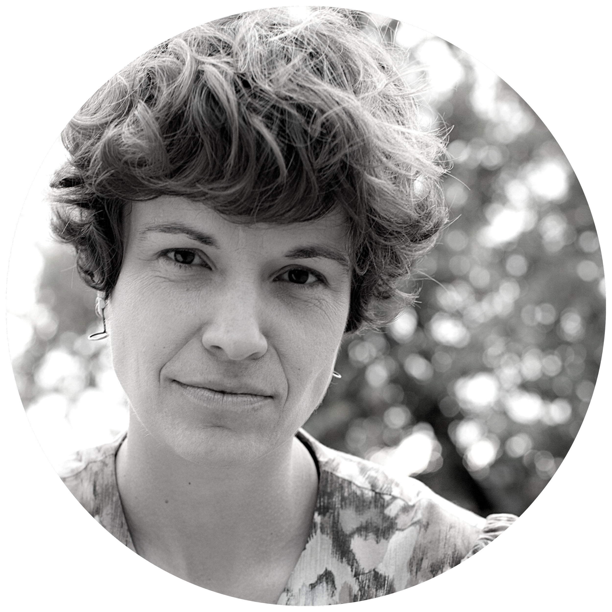 Black and white portrait of a woman with short, curly hair looking directly at the camera outdoors with a blurred background of trees.