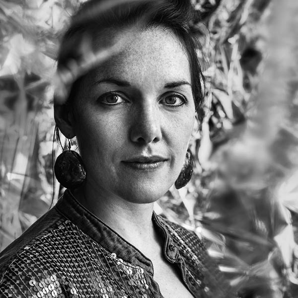 Black and white portrait of a woman with light skin, curly hair, wearing large earrings and a textured top, standing outdoors with foliage in the background.