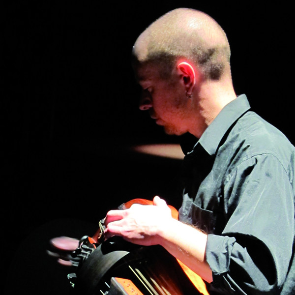 A person with blonde hair and earrings, dressed in a dark shirt, is looking down at an object in his hands against a dark background.