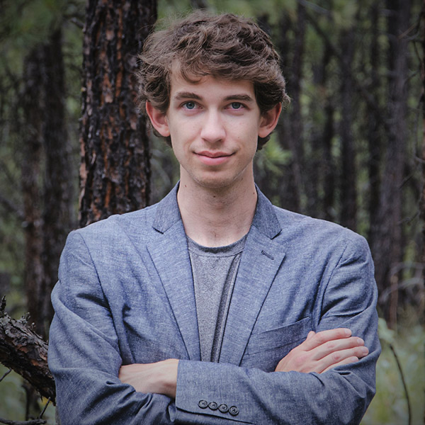 A young man with curly brown hair and blue eyes standing outdoors in a forest, wearing a gray blazer over a gray shirt, with his arms crossed.