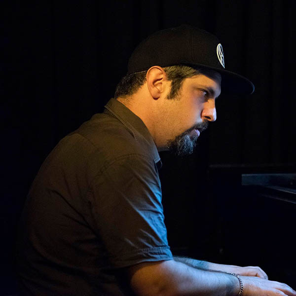A man with a beard, wearing a black cap and black shirt, playing a piano against a dark background.