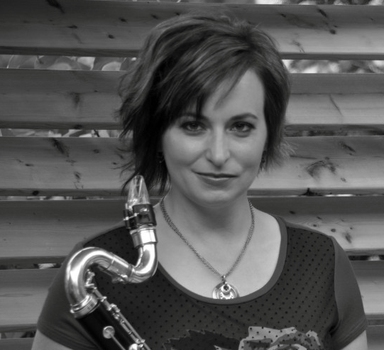 A woman with short, wavy hair holding a saxophone, standing in front of a wooden fence.