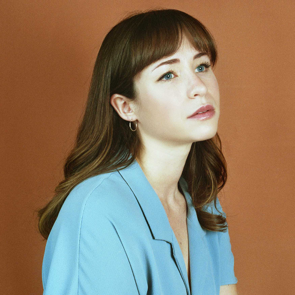 Portrait of a young woman with light skin, brown hair, and blue eyes, wearing a light blue shirt against a brown background.
