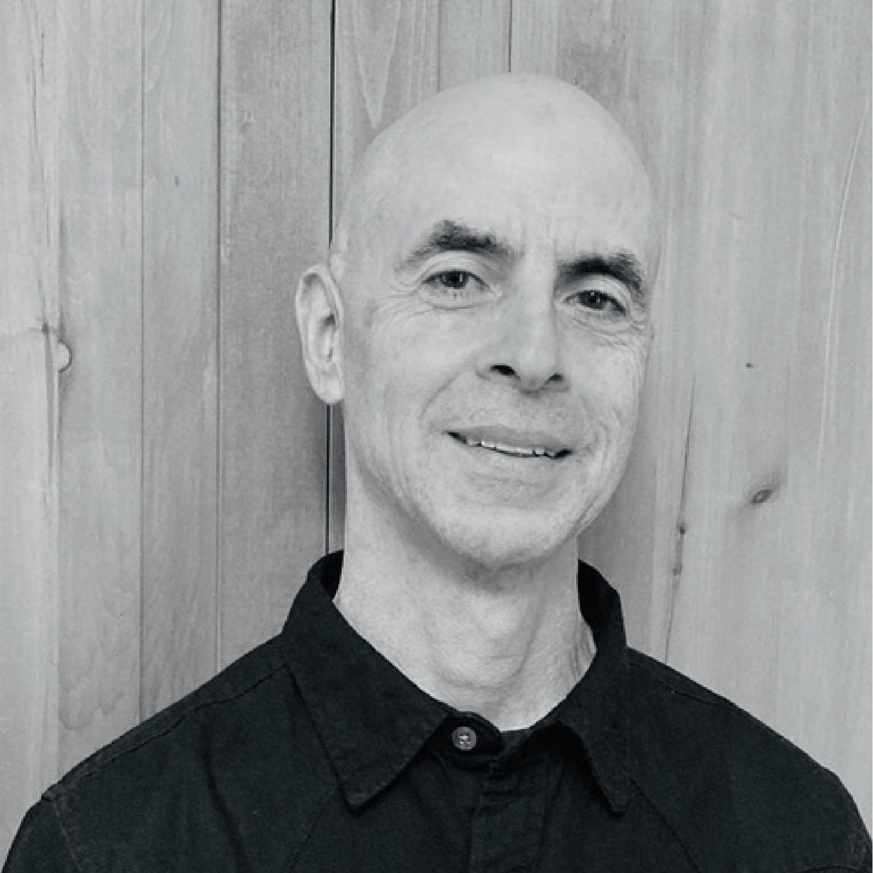 Black and white portrait of a smiling bald man wearing a black collared shirt, standing in front of a wooden wall