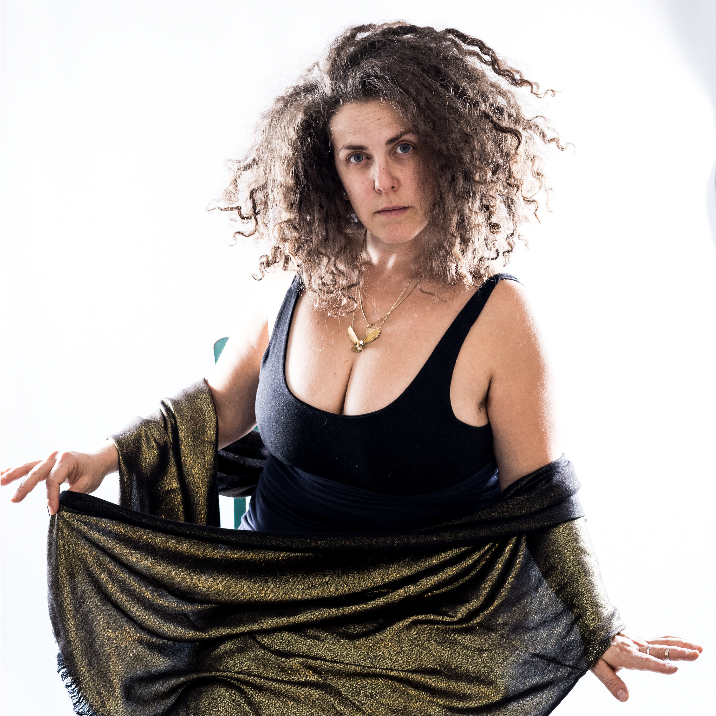 A woman with curly hair wearing a black top and gold skirt, holding the skirt out to the sides, against a plain white background.