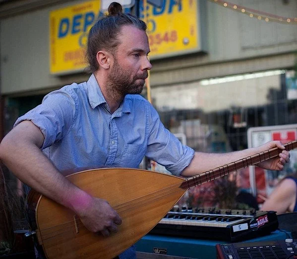 A man with a beard and long hair in a bun playing a stringed instrument with a keyboard in front of him.