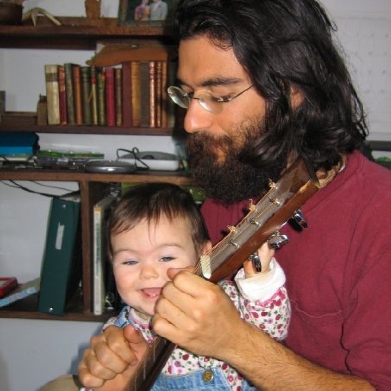 A man with glasses and long dark hair playing a guitar with a young girl smiling, holding the guitar's neck, in a room with bookshelves in the background.