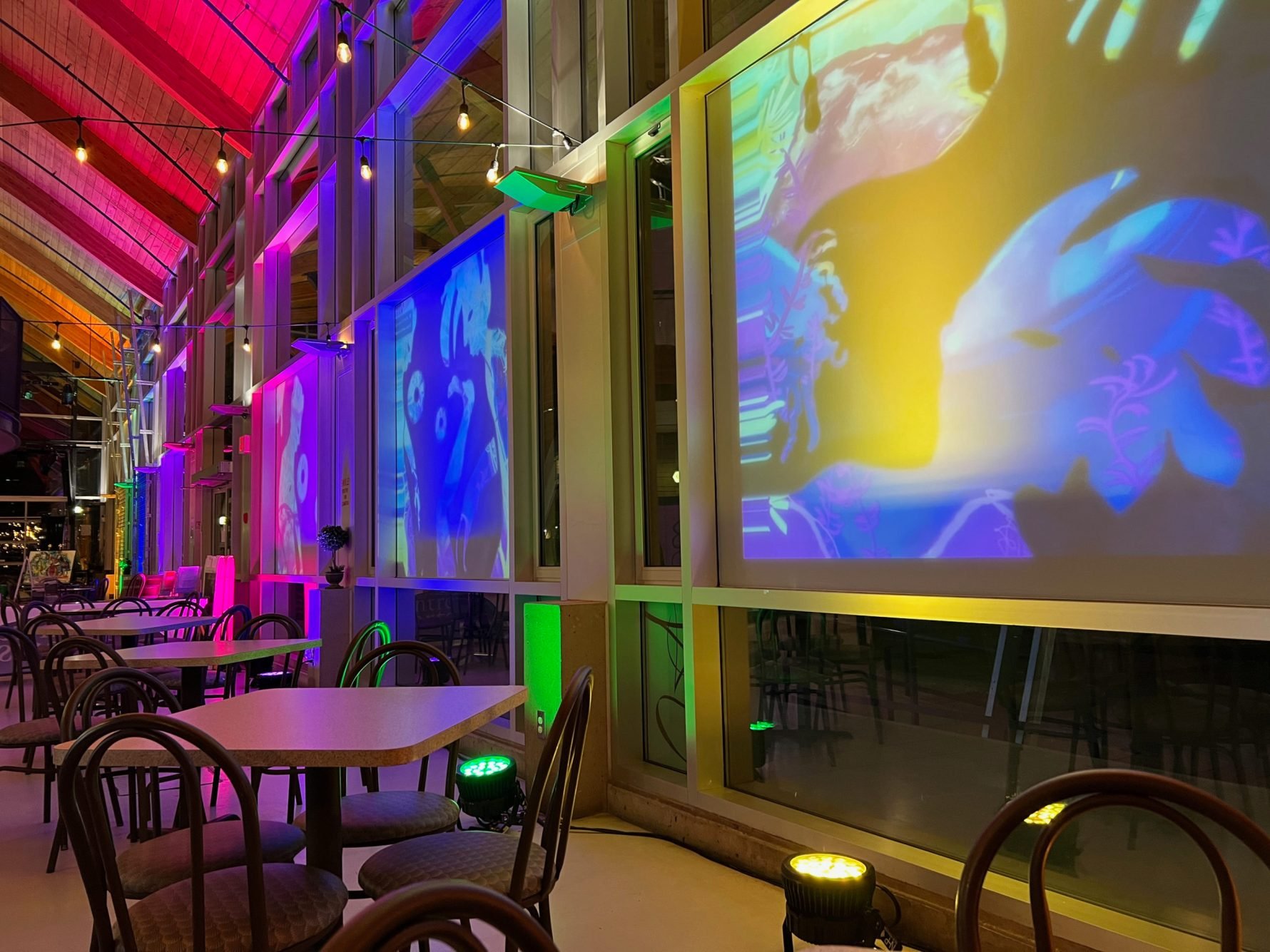 Colorful indoor restaurant or event space with illuminated wall projections, string lights, and tables with chairs.