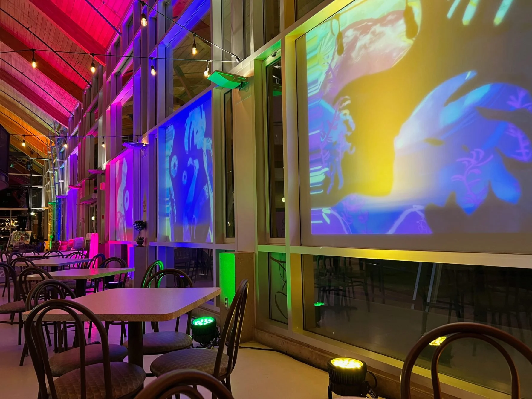 Indoor restaurant or cafe with colorful neon and LED lighting, large projection screens on windows displaying abstract and floral visuals, and empty tables and chairs.