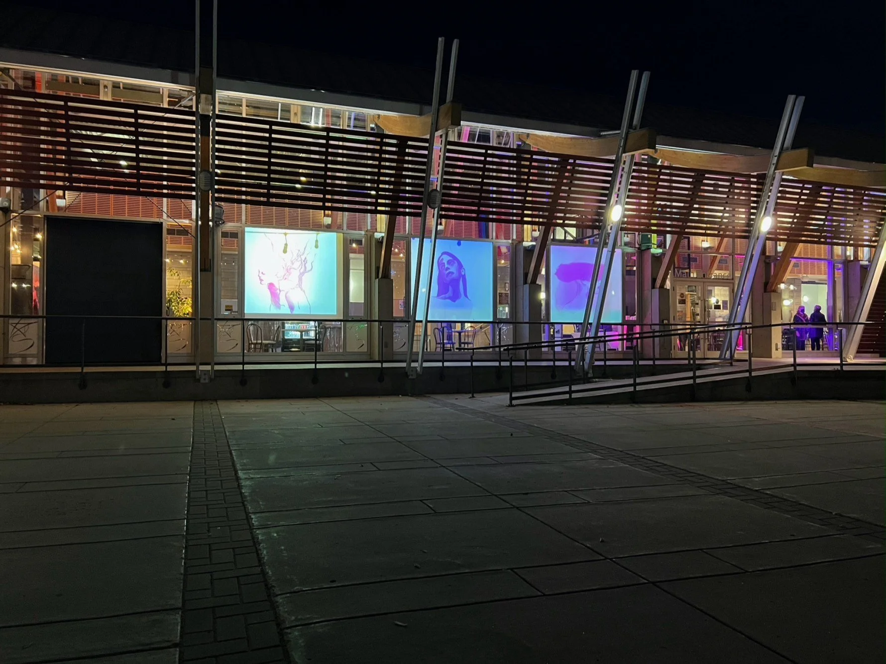 Night view of a modern building with large glass windows displaying colorful digital artwork and interior lights. Exterior appears empty with a paved walkway in front.