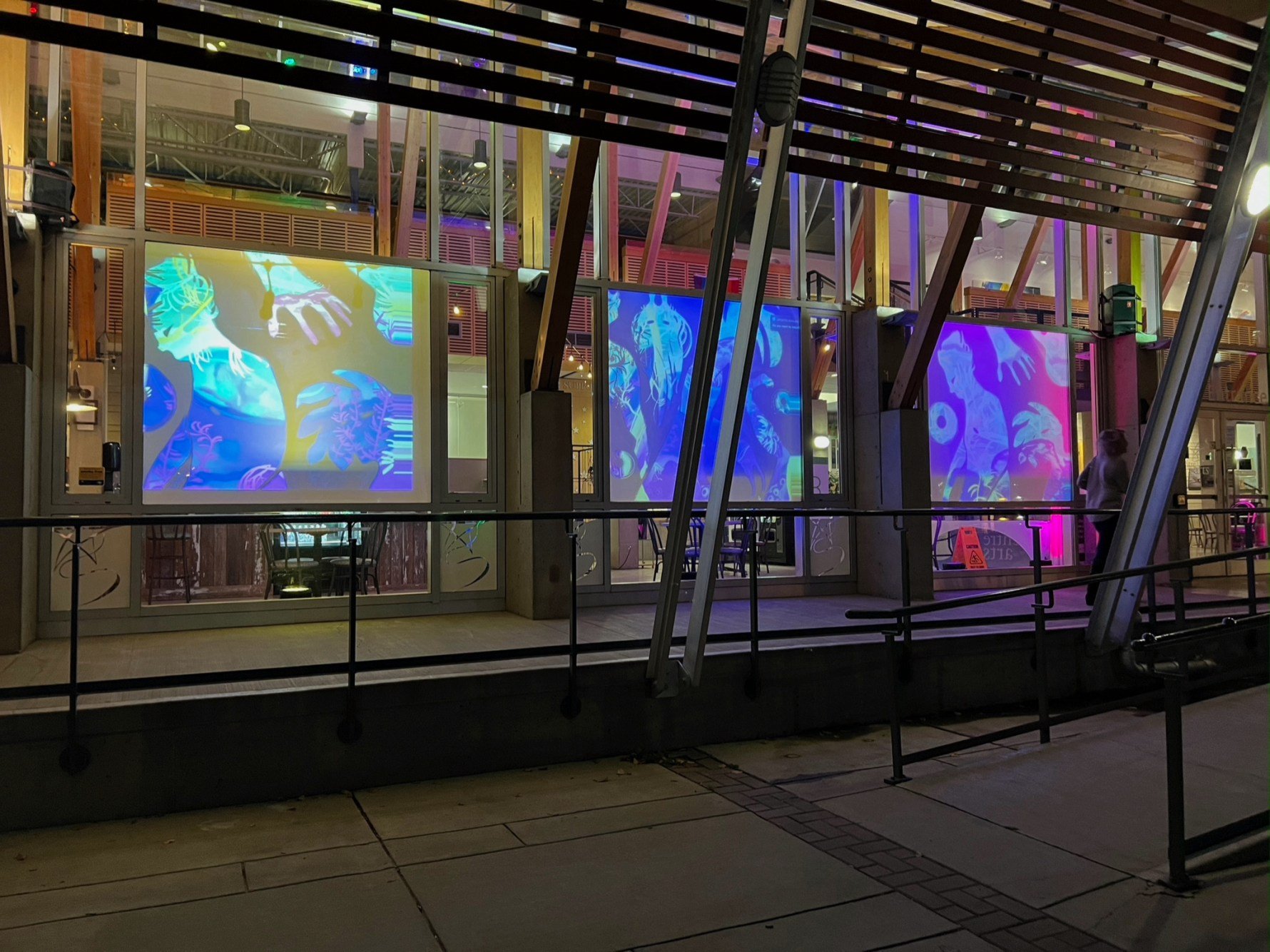 Nighttime view of a modern building with large glass windows displaying colorful, abstract, multi-colored digital art projections. The building has an architectural design with angled beams and wooden accents. A person is standing outside the buildin