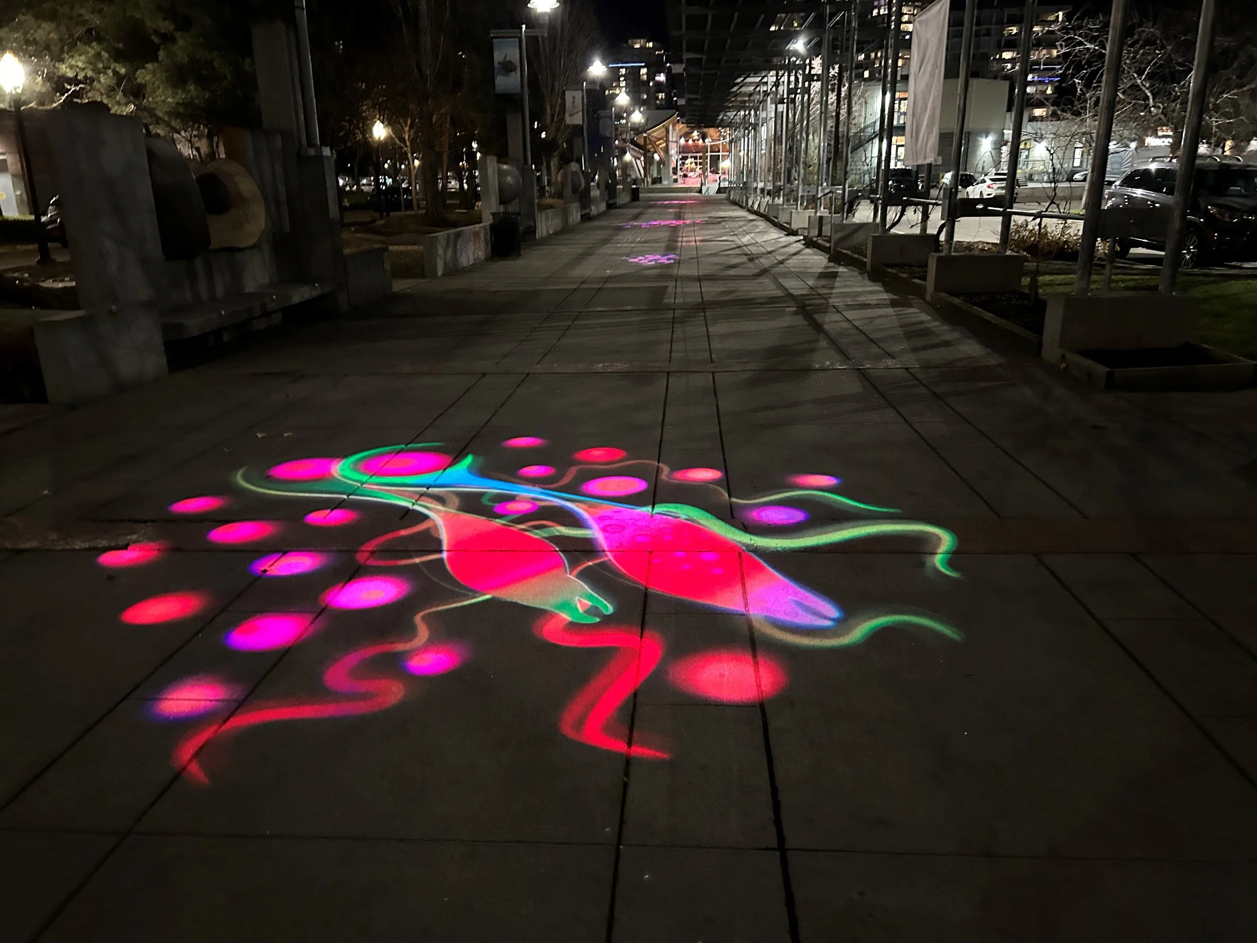 Colorful stenciled sidewalk art of a pink and green dragonfly with pink dots, illuminated at night.