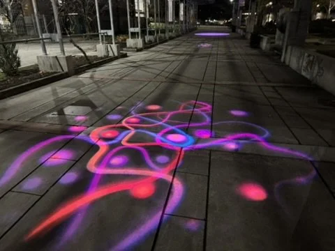 Nighttime sidewalk with colorful neon laser light projections creating abstract patterns.