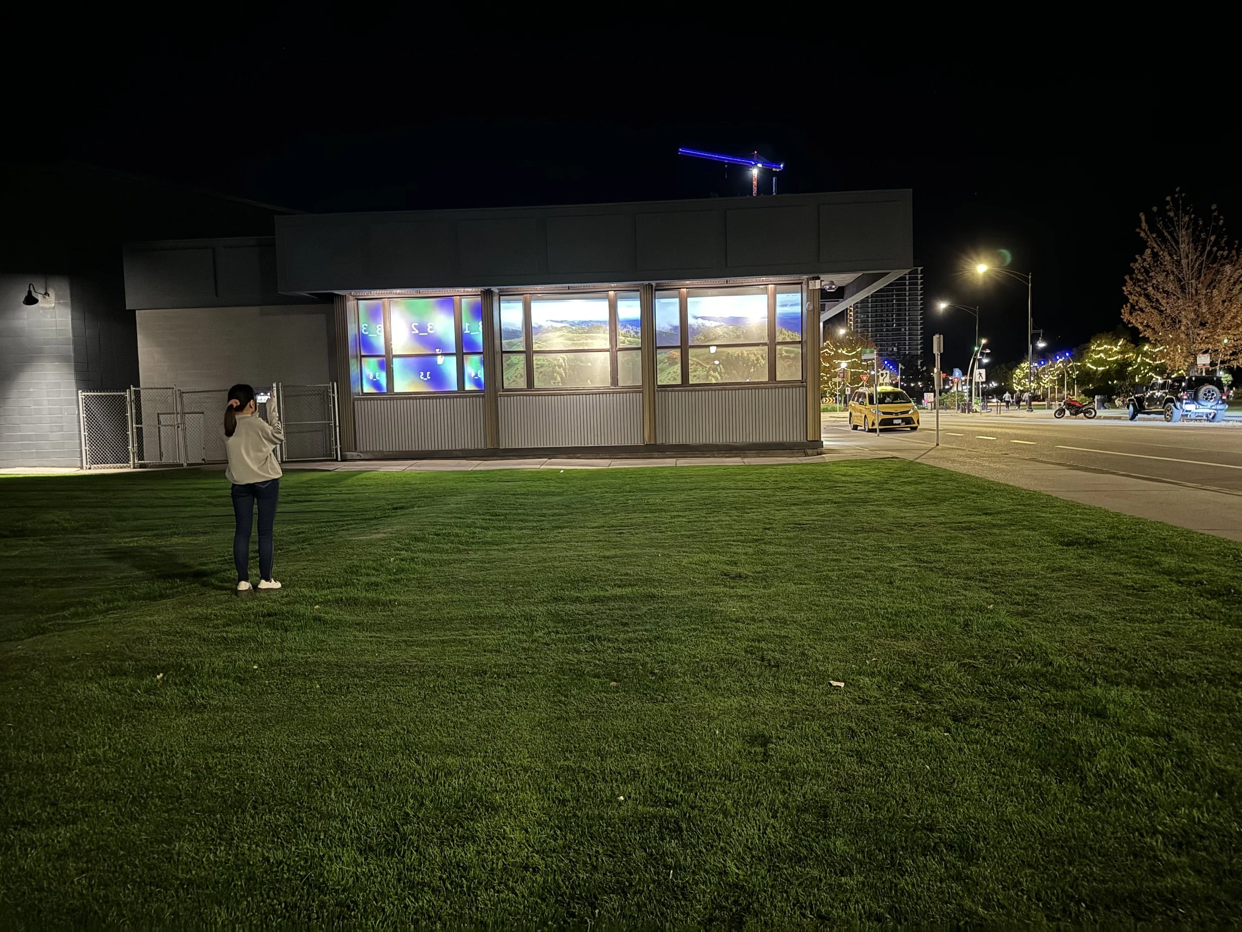 A woman standing on a grassy area taking a photo of a building at night. The building has illuminated windows displaying landscape images, and there are lights and vehicles along a nearby street.