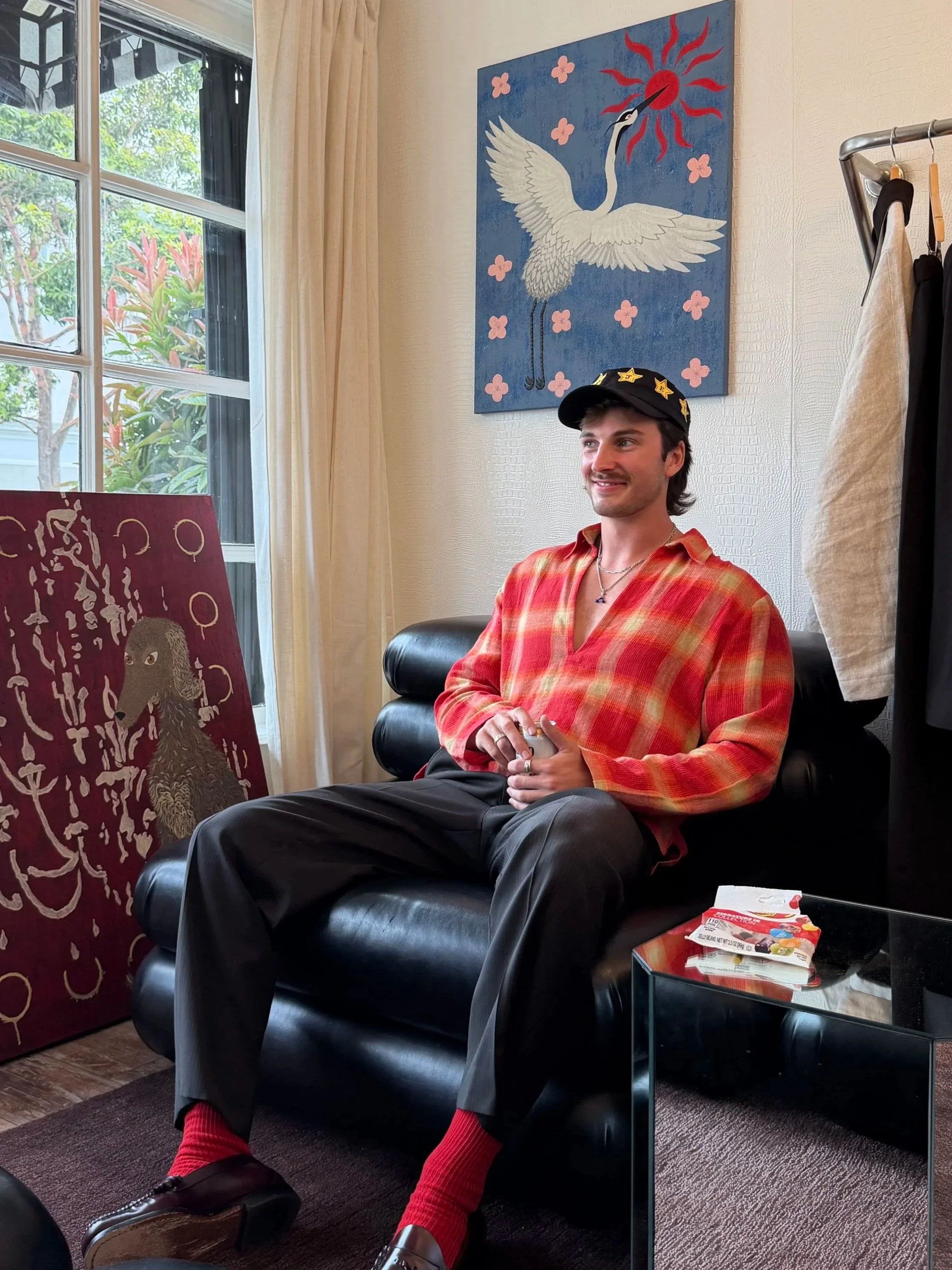 A young man with dark hair, a mustache, and a beard sitting on a black leather chair in a room with large windows. He is wearing a black cap with yellow stars, a red and orange plaid shirt, black pants, red socks, and black shoes. Behind him, there is a painting of a white crane with outstretched wings on a blue background with pink flowers. To his right, there is a small table with a colorful box, a stand with hanging clothes, and a window with beige curtains and outside greenery.