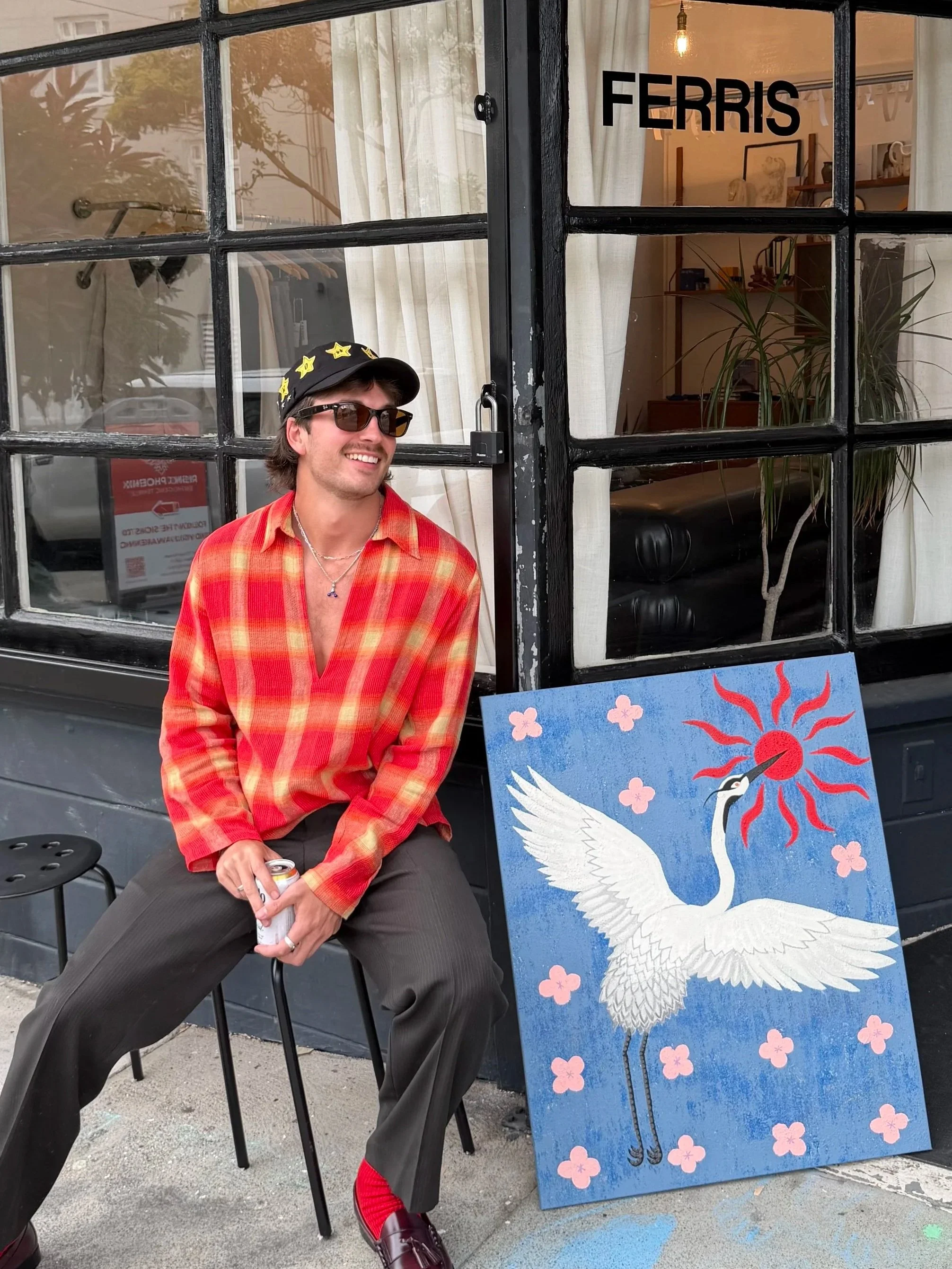 A man wearing sunglasses, a black cap with yellow stars, a red plaid shirt, and gray pants, holding a soda can, sitting on a black chair outside a shop named 'FERRIS'. There is a colorful painting of a white crane with a red sun and pink flowers on an easel next to him.