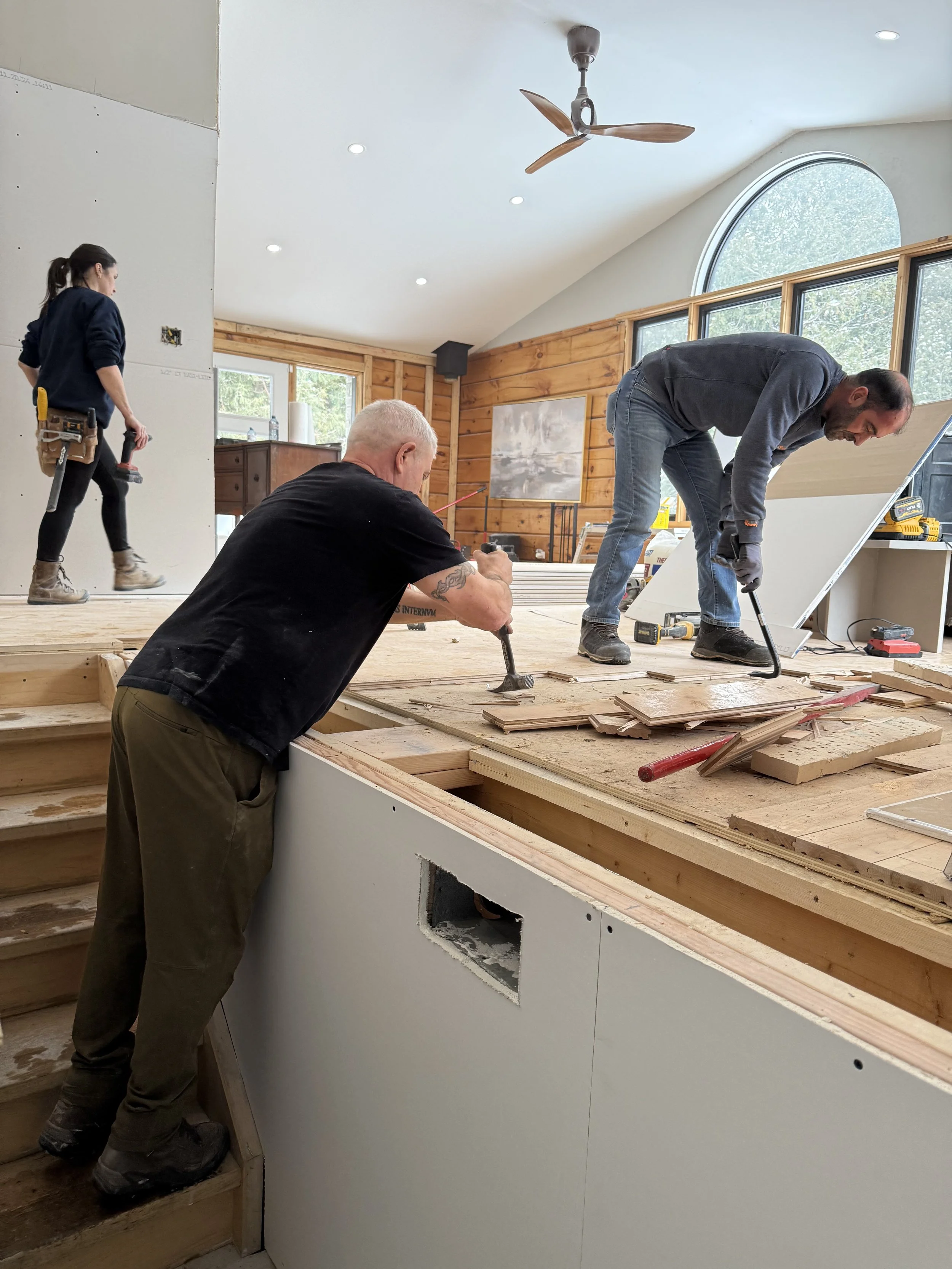 People installing wooden flooring in a room with wood-paneled walls and large arched windows.