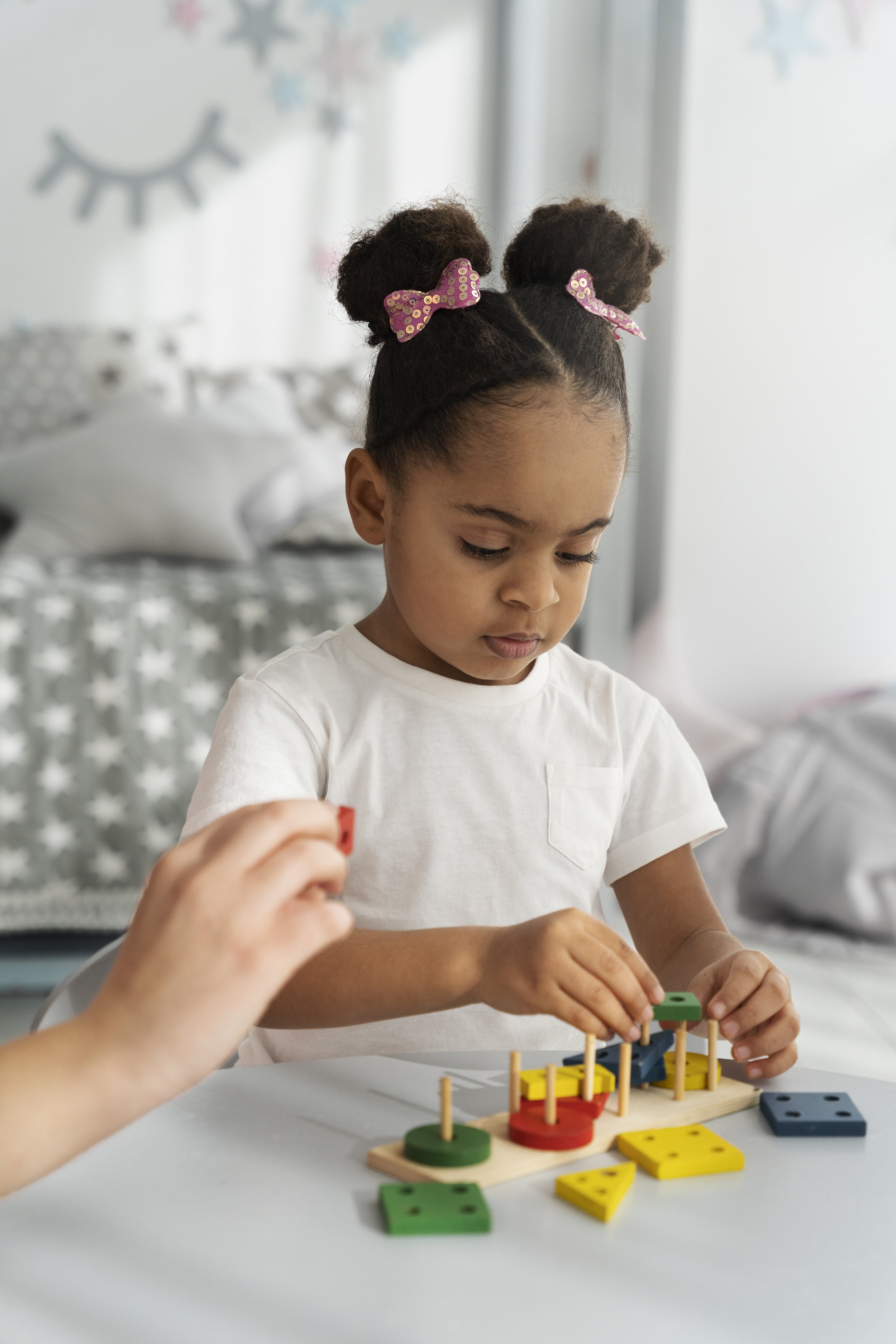 A young girl with curly hair styled in two buns secured with pink polka dot bows is playing a colorful wooden logic game. The game pieces are various shapes and colors, including yellow, green, and blue, on a white table in a well-lit room with star-patterned bedding and festive decorations.