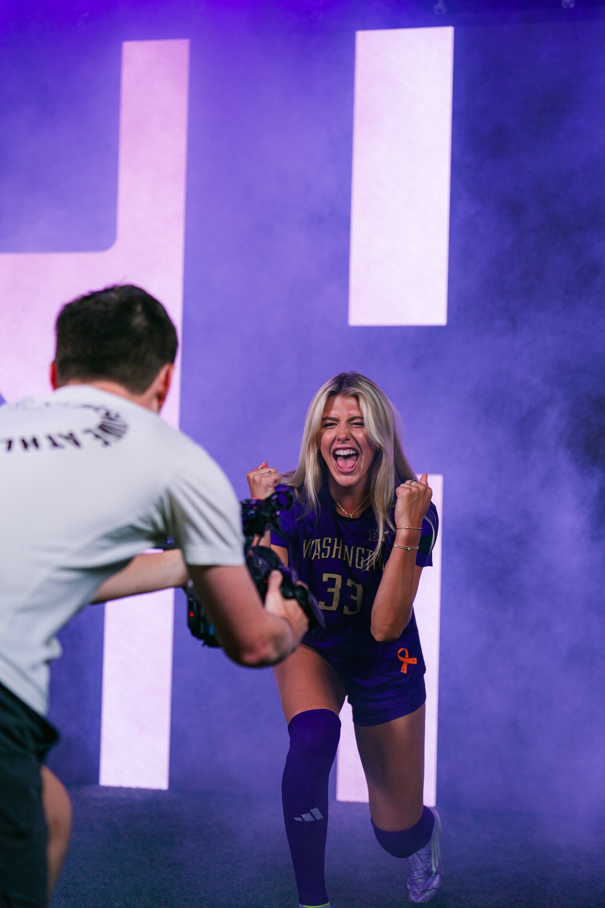 A young woman in a Washington soccer  jersey is celebrating with a joyful expression, fists clenched, during a sports event. A photographer is capturing her moment.