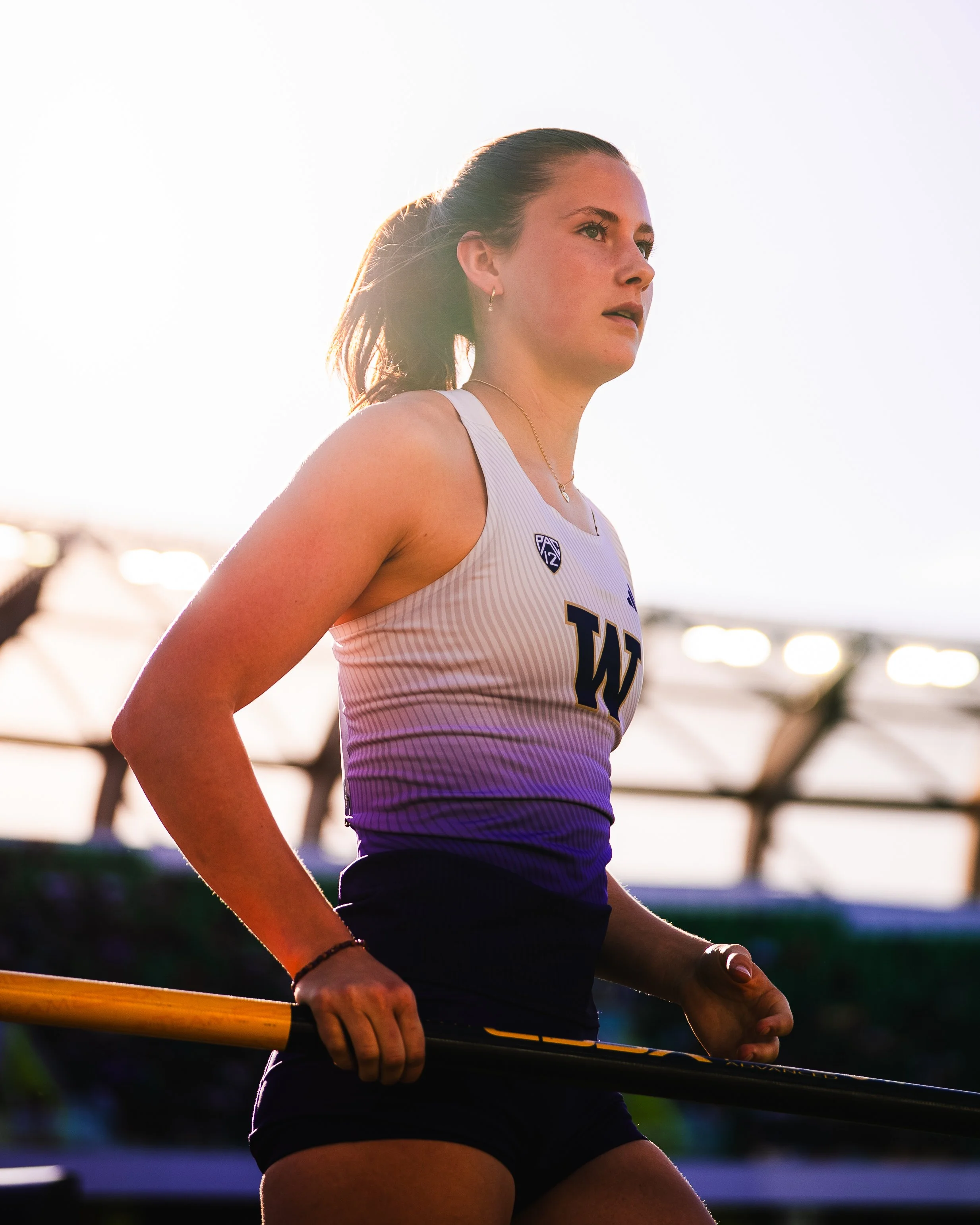 A female athlete in a white sports tank top with UW logo, holding a yellow pole vault pole in her right hand, standing outdoors during sunset.