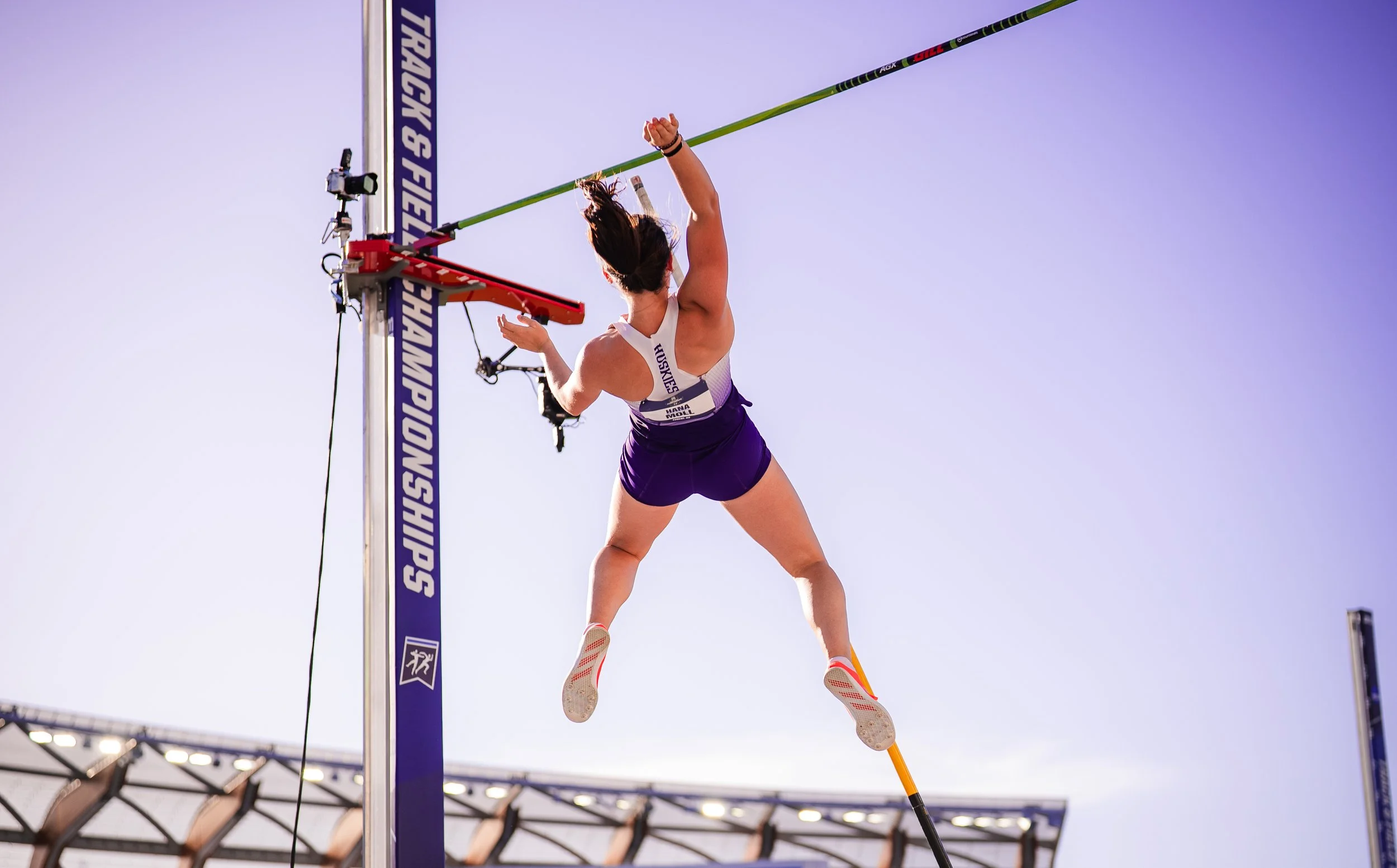 A female athlete in a purple sports uniform competing in hammer throw during a track and field event, with a large stadium and blue sky in the background.