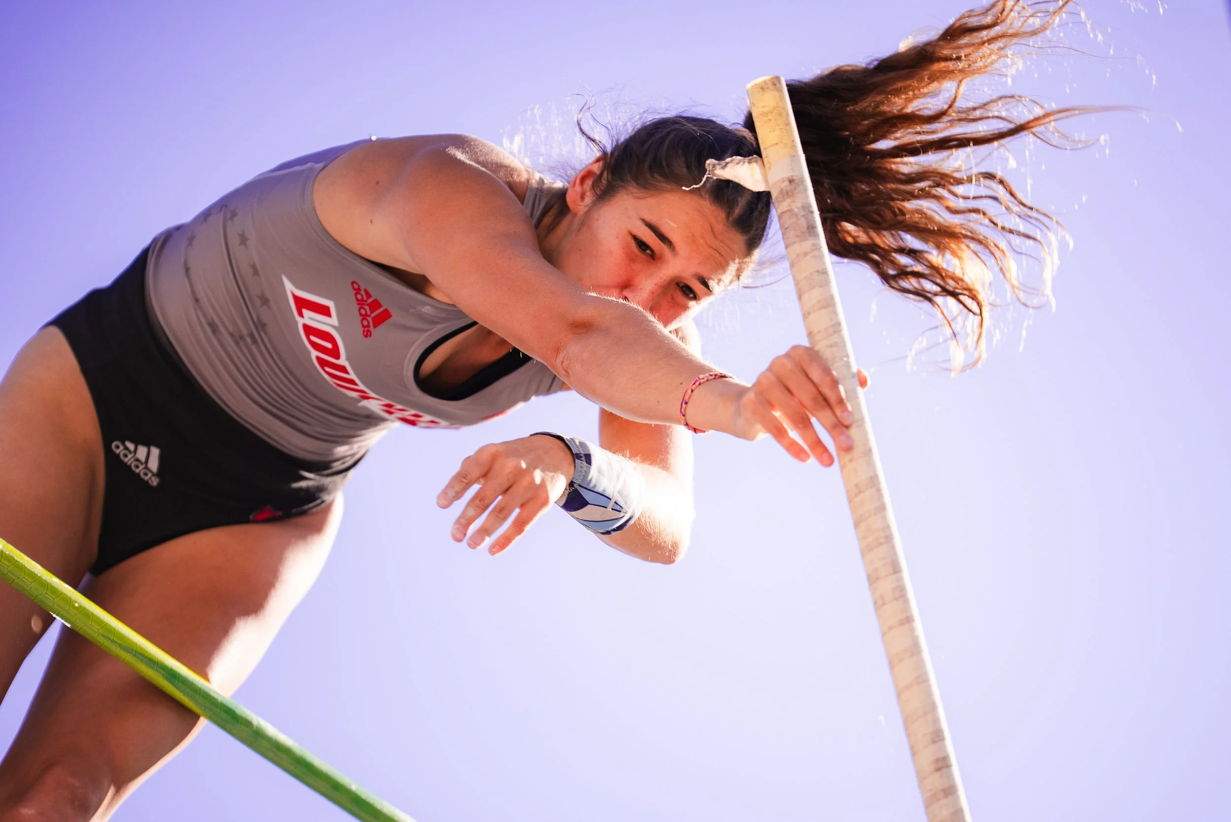 A female pole vaulter in a gray Adidas athletic top and black shorts preparing to clear a high bar during a track and field event, with clear blue sky in the background.