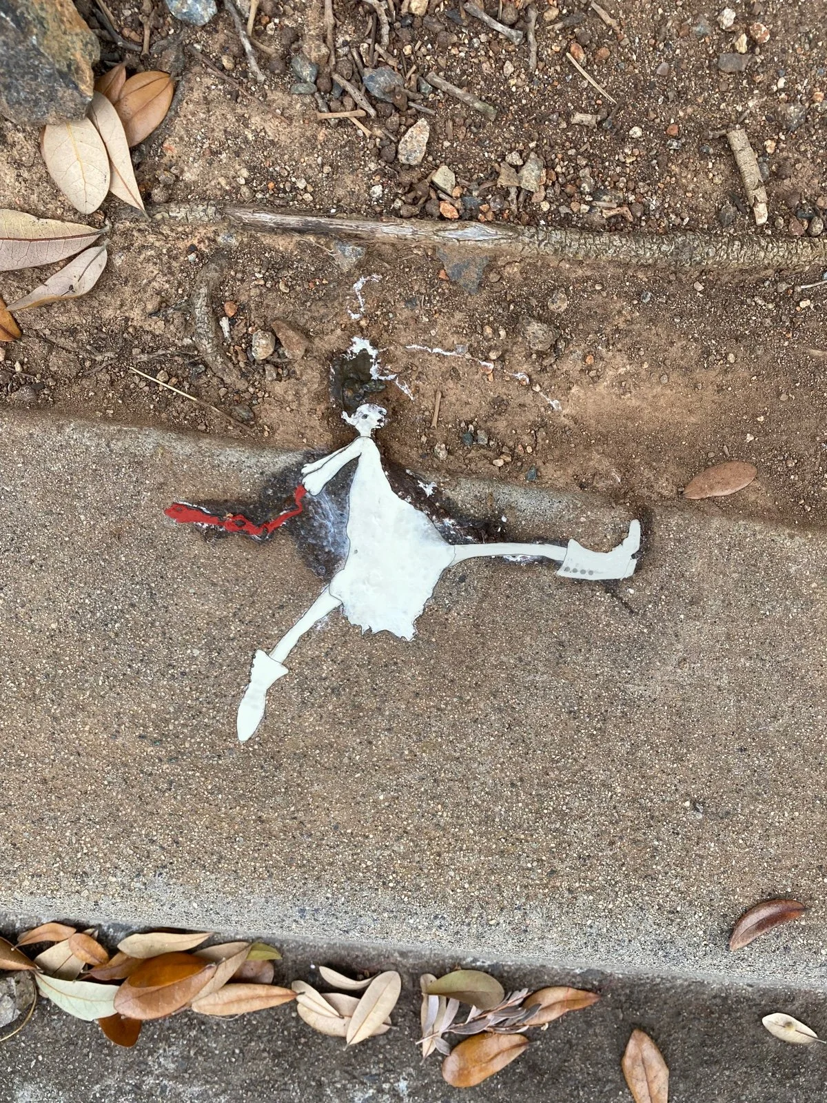 A torn paper figure of a girl with a white dress and white boots lies on the ground next to a curb, with dry leaves scattered around.