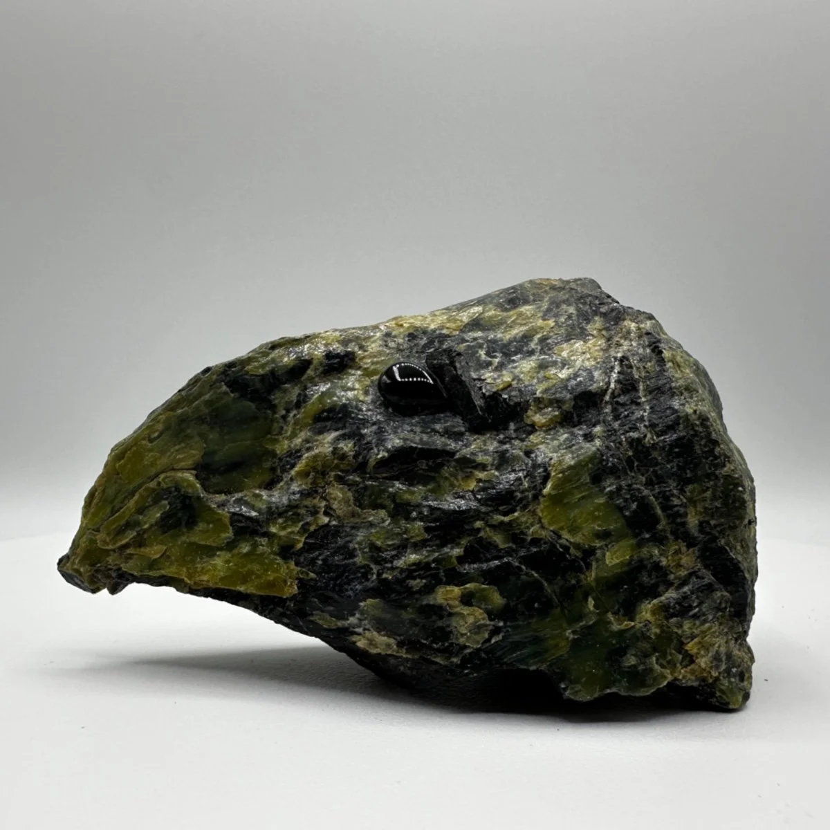 A rough, dark green and black rock with a glossy, black obsidian fragment on top, against a plain gray background.