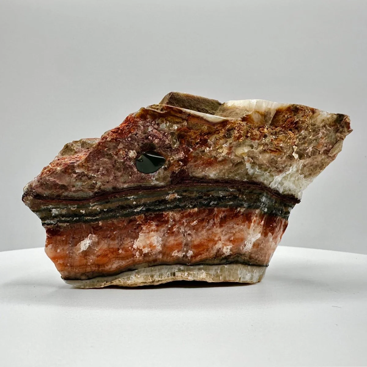 A polished mineral specimen with layered bands of reddish, white, black, and gray minerals, featuring a black spot, placed on a plain white surface against a light gray background.