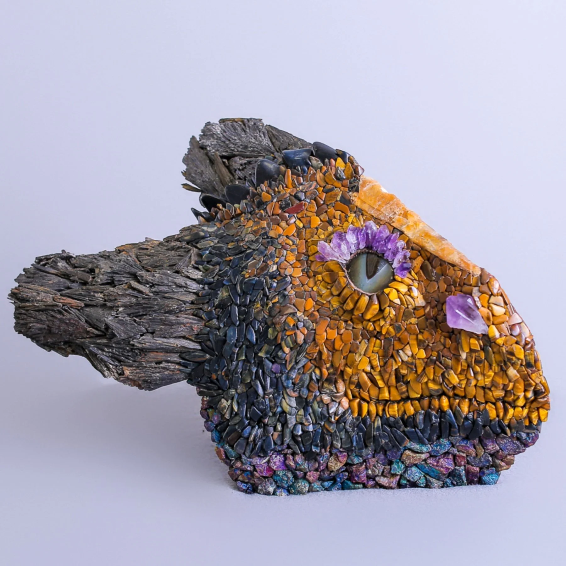 A mosaic art piece shaped like a fish, composed of colored stones, shells, and gems, with an eye made of a large shell and purple gemstones, set against a plain background.