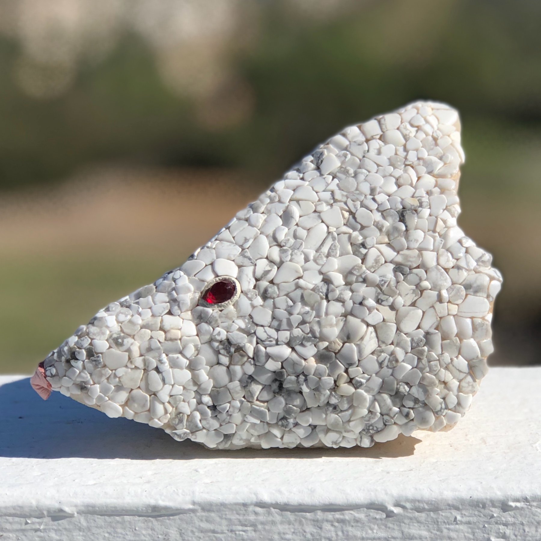 A close-up of a small beaded gecko figurine with red eyes, made of white and gray stones, resting on a white painted surface outdoors.