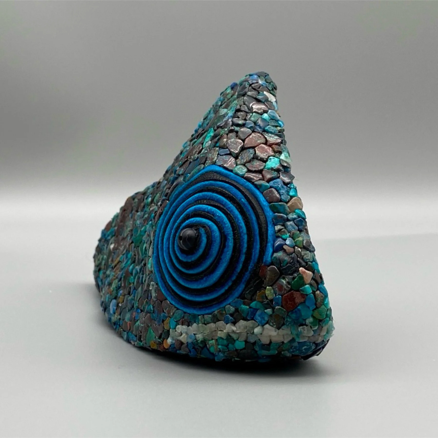 A decorative, cone-shaped object covered in multicolored small stones and featuring concentric blue rings on the front, set against a plain gray background.