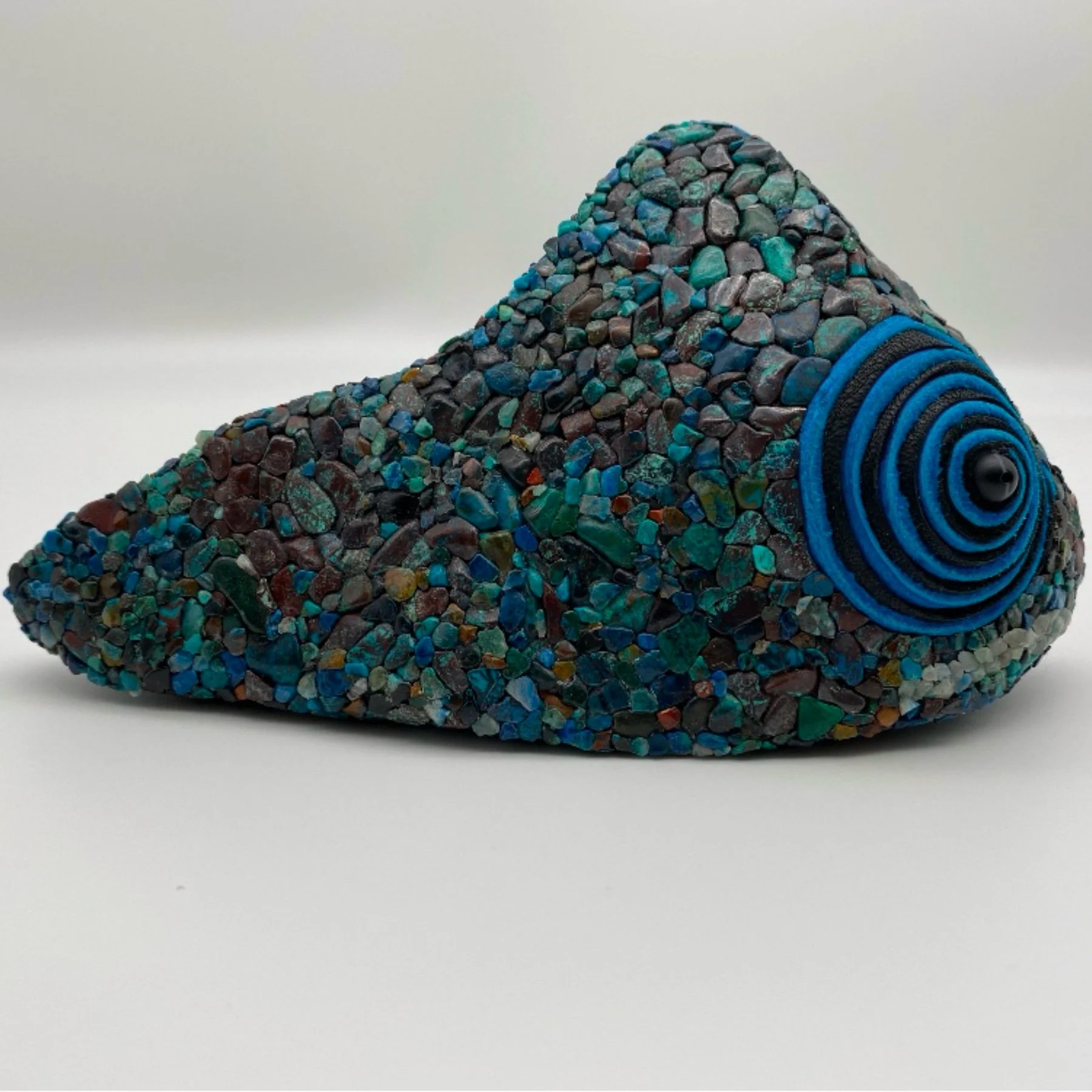 Zoomed in view of a multicolored pebble-covered rock with a blue and black spiral pattern on the right side, placed on a white surface.