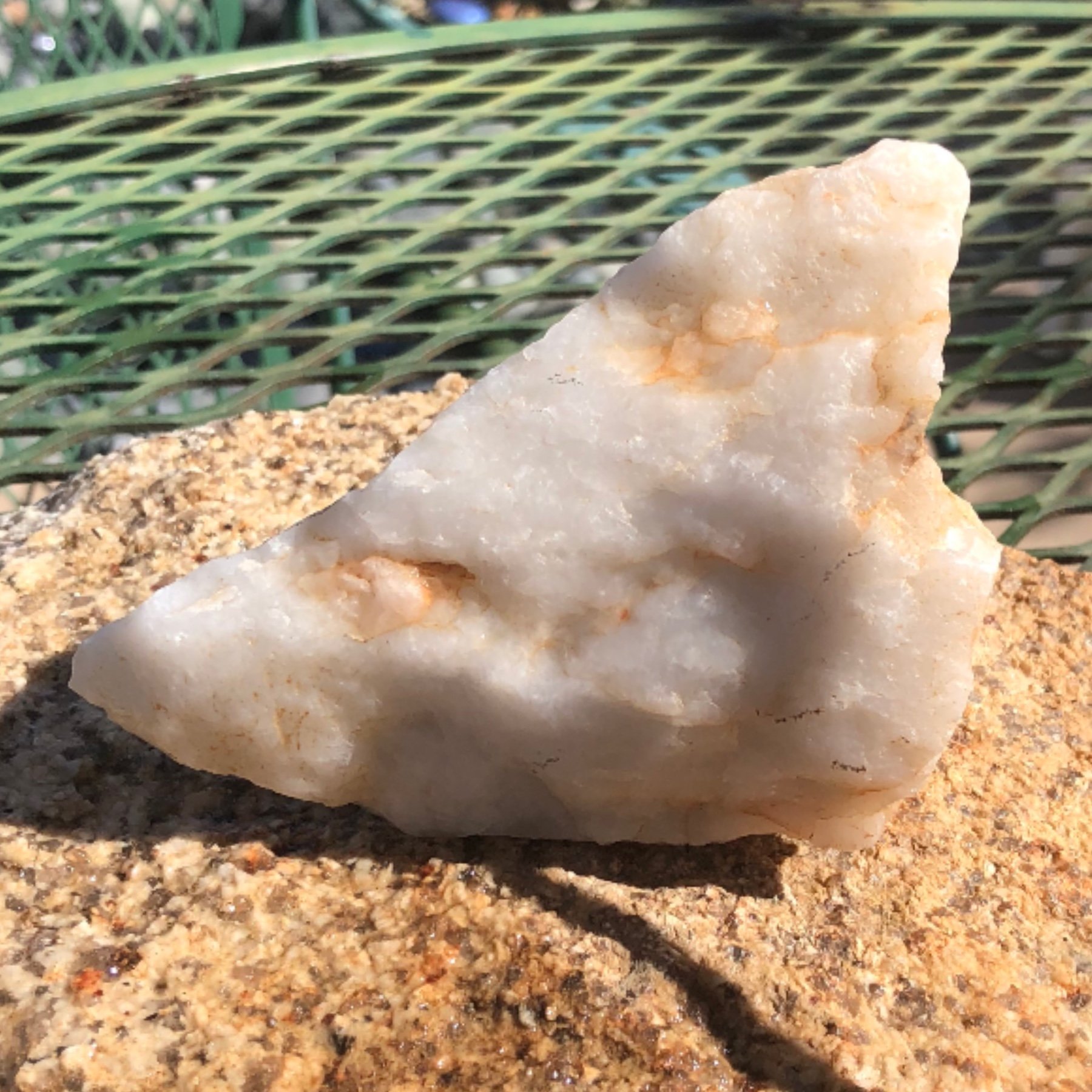Close-up of a rough, white and orange mineral rock on a speckled granite surface with a green wire fence in the background.