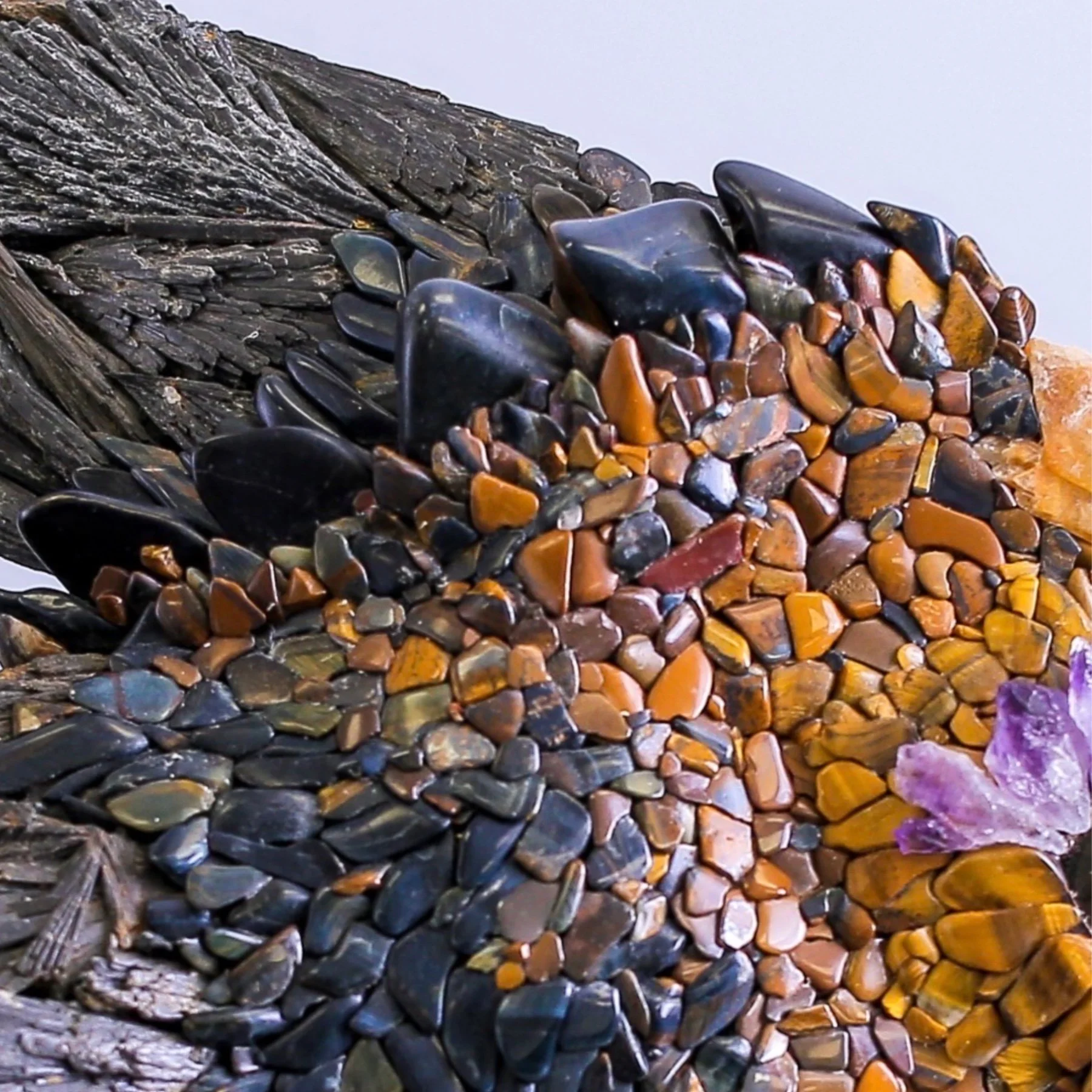 Close-up view of a layered rock sculpture made of polished stones of various colors, with purple flowers visible on the bottom right.