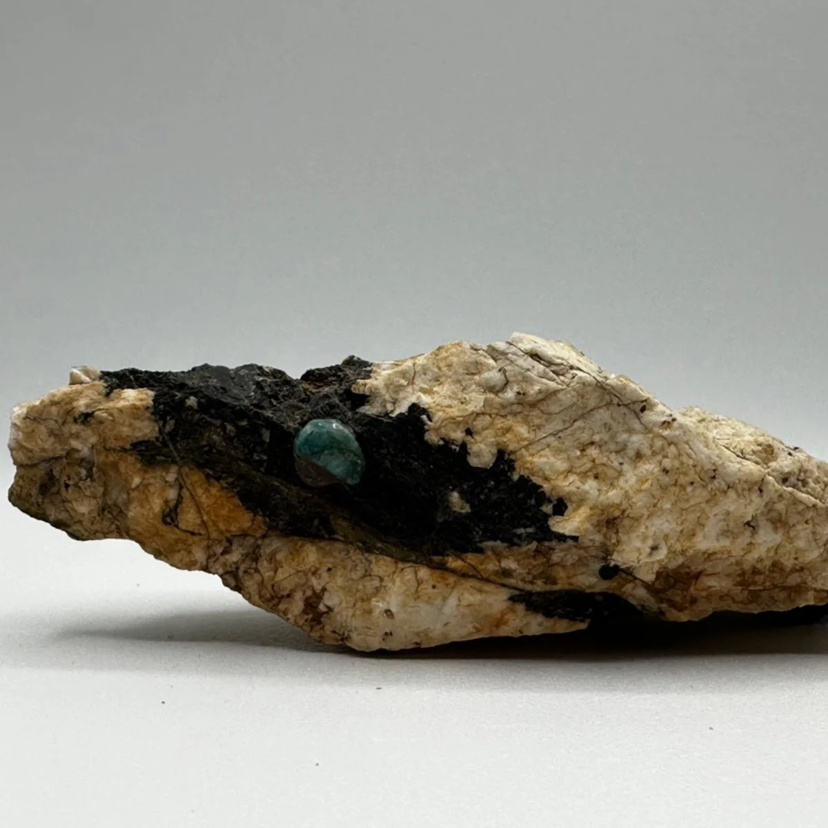 A rock with black material and a small greenish-blue mineral or gemstone embedded in it, placed on a plain surface with a neutral background.