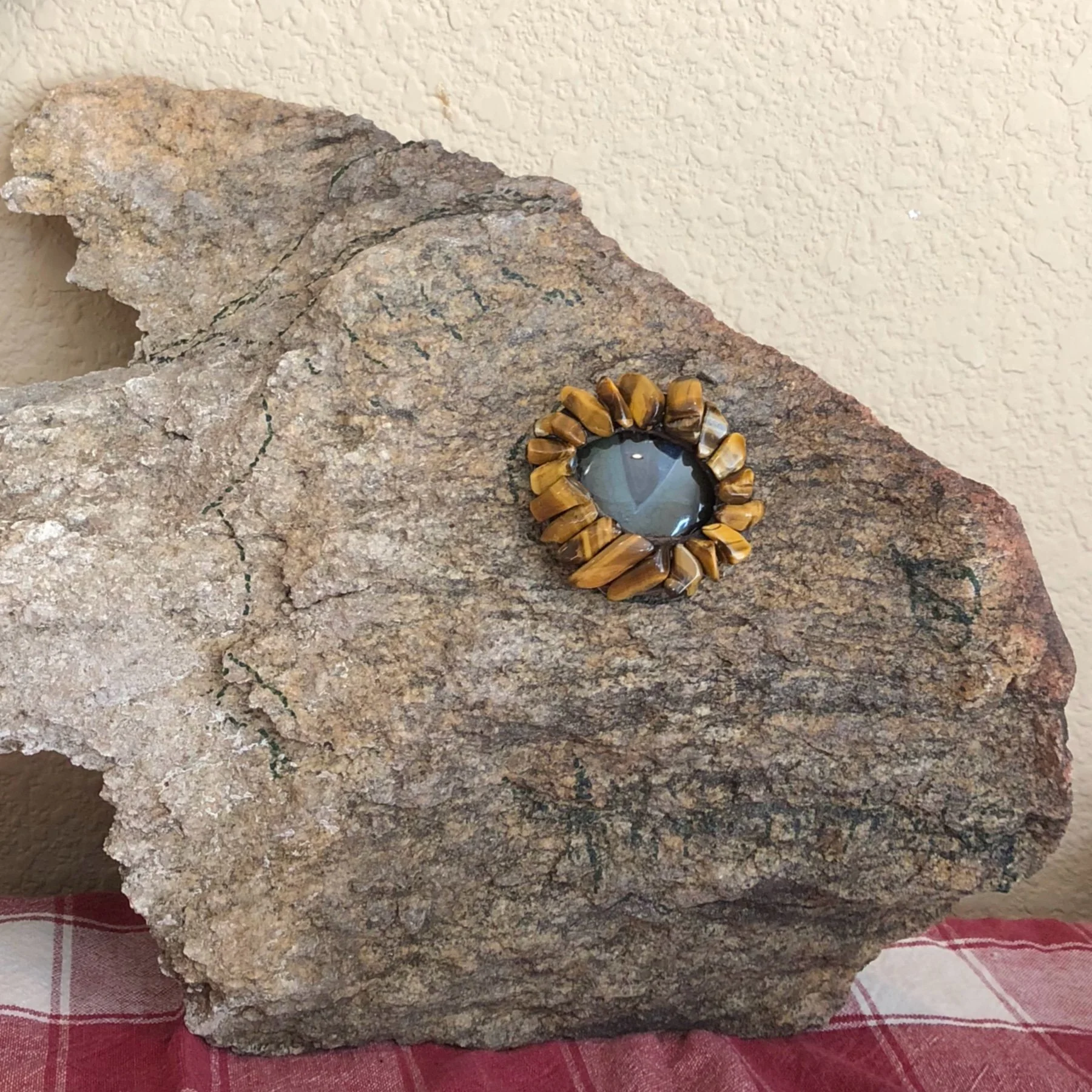 A large piece of rough granite rock with a round, decorative mirror attached to it. The mirror has a border made of tiger's eye stones. The background features a textured beige wall and a red and white checkered cloth underneath the rock.