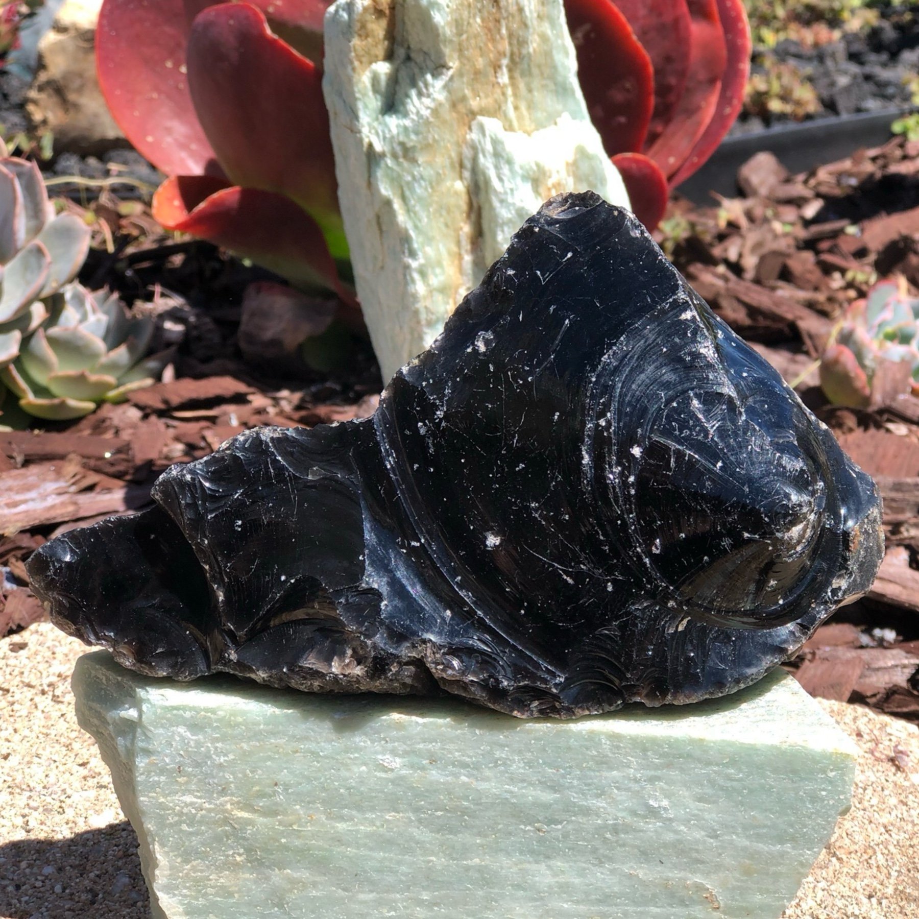 Close-up of a rock sculpture composed of several stones, including a large black piece with white streaks at the forefront, a beige and green stone behind it, and red succulents on the left and background, set against soil and mulch.