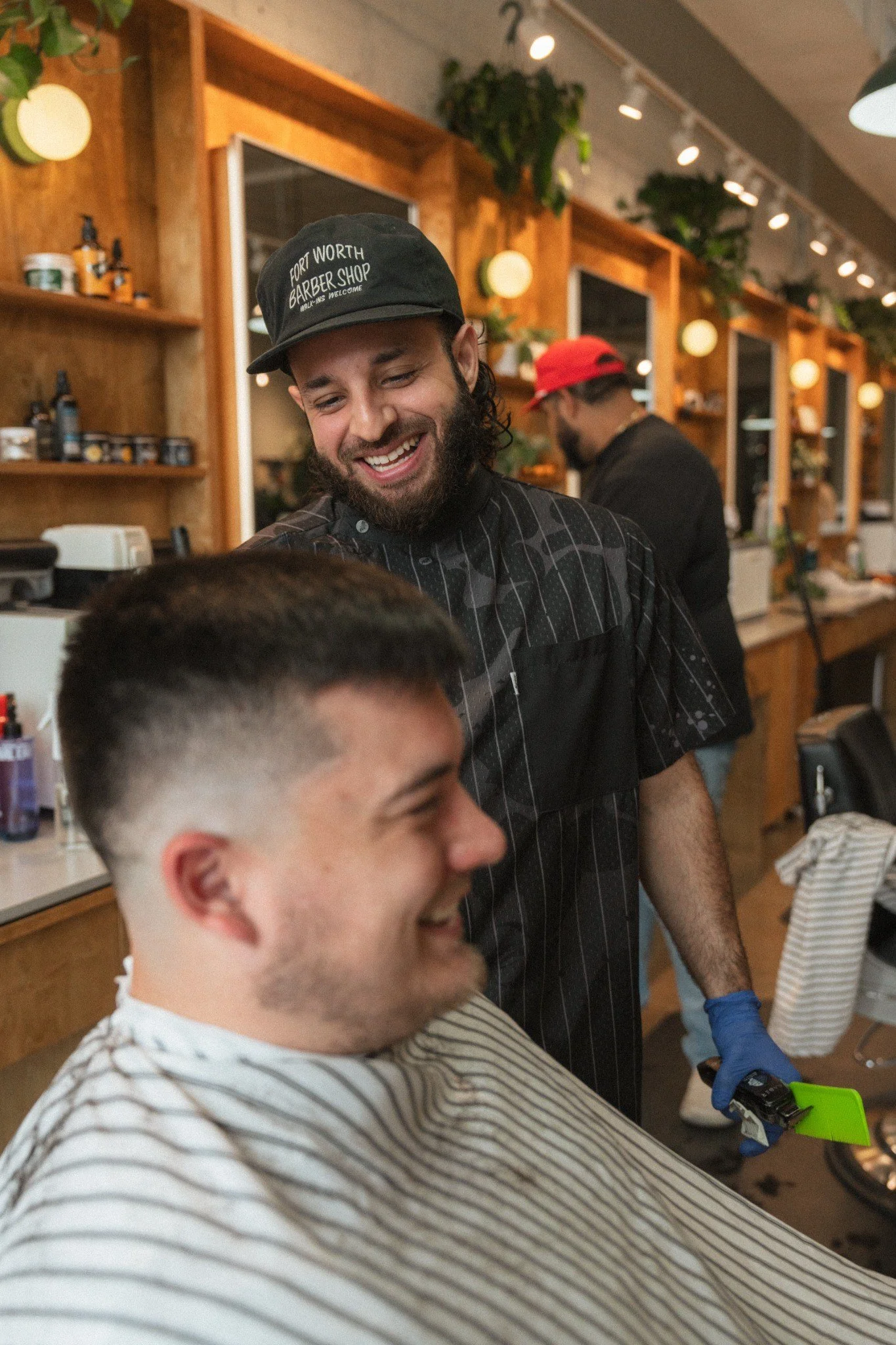 A man with a beard and a black cap smiling at a barber shop while getting a haircut. The barber shop has wooden shelves, mirrors, and plants. Another man is in the background wearing a red cap.