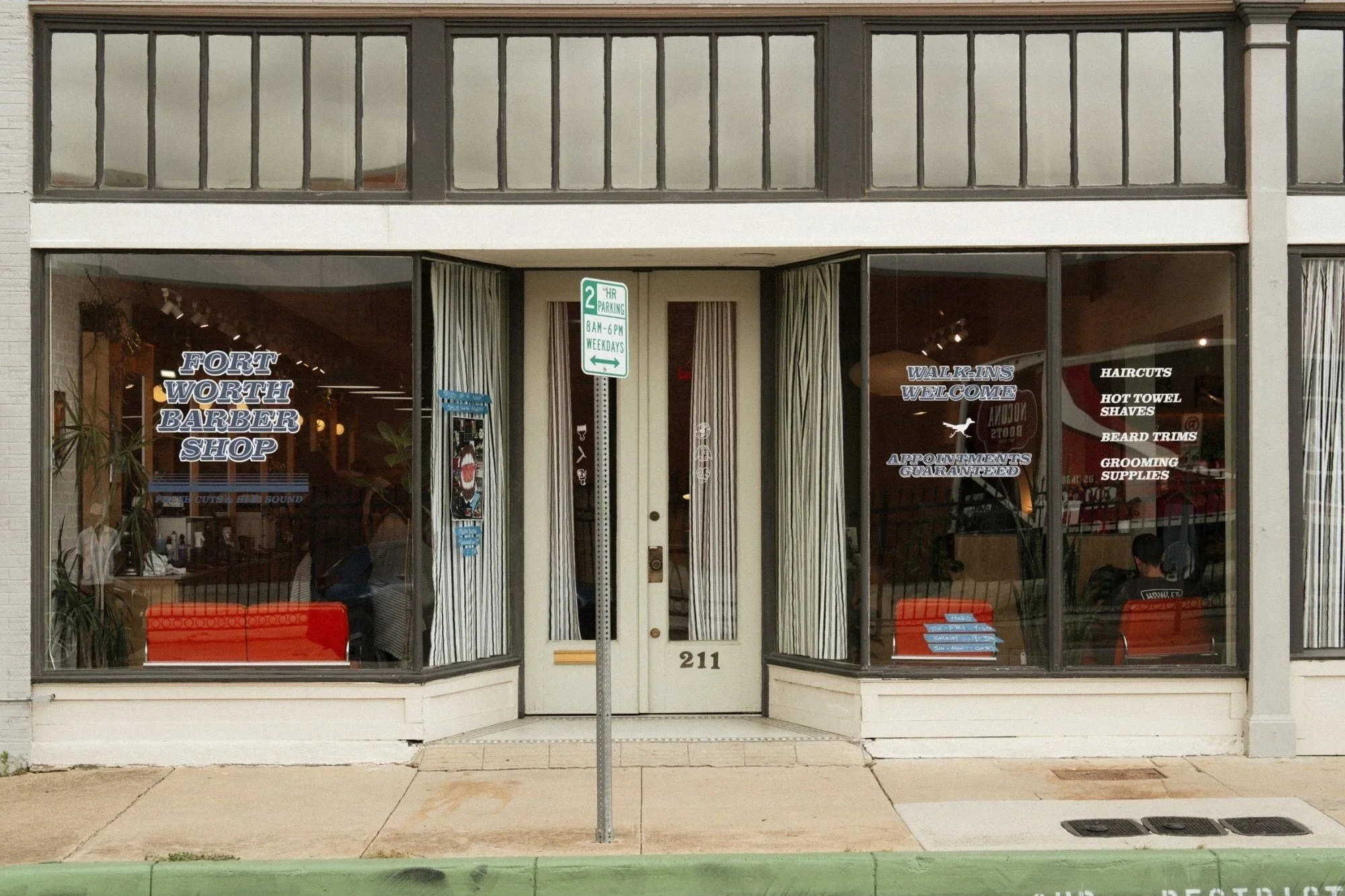 The storefront of Fort Worth Barber Shop with large glass windows and a glass door. The shop has signs on the windows indicating services such as walk-ins welcome, haircuts, hot towel shaves, beard trims, grooming supplies, and appointment guarantees. A parking sign in front indicates 2-hour parking from 8 AM to 6 PM on weekdays. Inside, there are people seated and waiting, and plants are visible near the left window.
